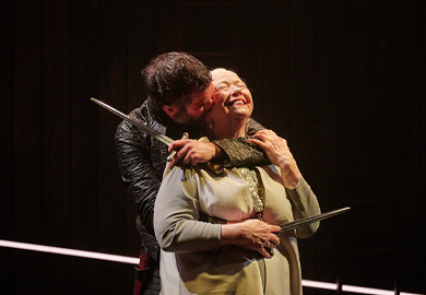 A tense stage moment from Macbeth featuring an actor holding a dagger to the throat of an older actress, who smiles with her eyes closed