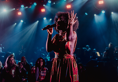 Alt text: An image of a person in a colorful dress and jewelry singing into a handheld mic. 