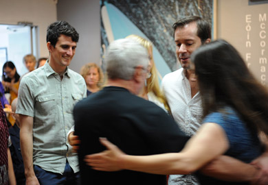 A group of people are dancing in a circle with their arms around each other on the right side of the photo. A person to their left smiles as they watch them.  